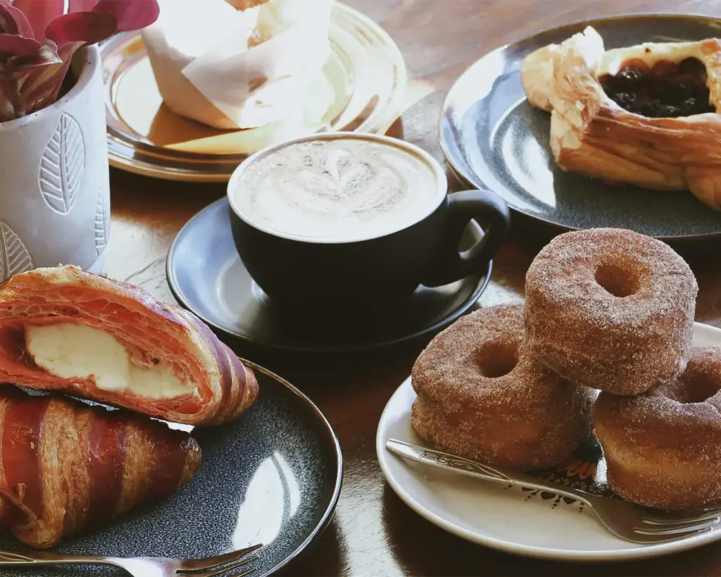 Assorted pastries including croissants, cinnamon sugar donuts, and a fruit danish, with a latte in a black cup on a wooden table.