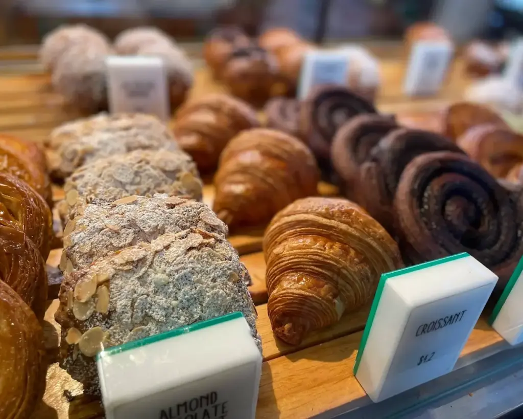 A variety of pastries on a bakery shelf, featuring almond croissants dusted with powdered sugar, flaky golden croissants, and chocolate swirls.
