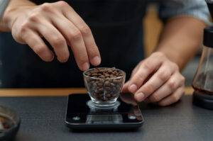 Close-up of a barista's hands weighing whole roasted specialty coffee beans in a small glass cup on a digital scale.