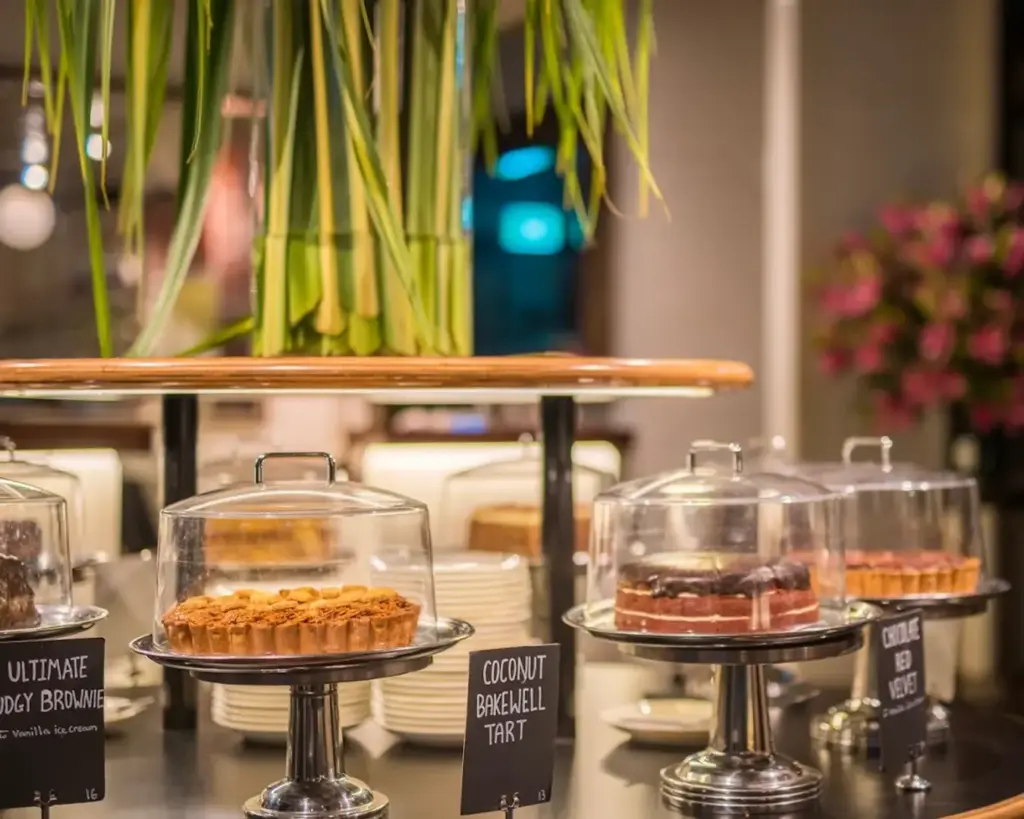 Assorted cakes and tarts displayed on stands with clear covers and labels in a cozy bakery setting.
