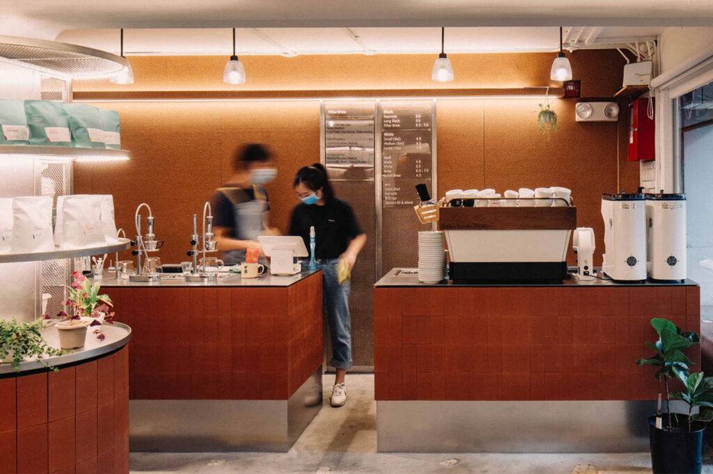 Interior view of a modern coffee shop counter with red tile and cork walls, featuring espresso machine and two baristas working.