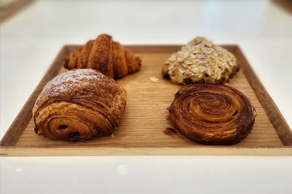 A selection of freshly baked pastries, including a pain au croissant, a classic croissant, a nut-topped croissant, and a spiral danish, served on a wooden tray at %Arabica.