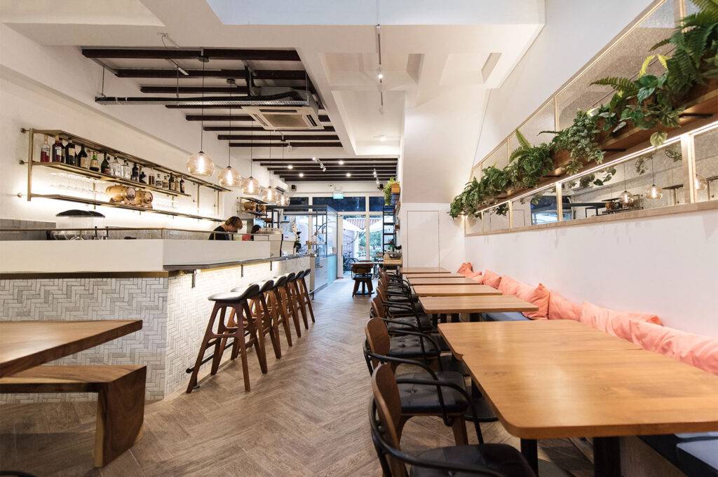 Bright, chic cafe interior with long wooden tables, black chairs, and a white bar. Potted plants line the mirrored wall shelf.