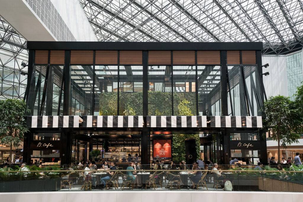 Modern, two-story glass facade cafe with a striped awning and a living green wall, located inside a large mall atrium.