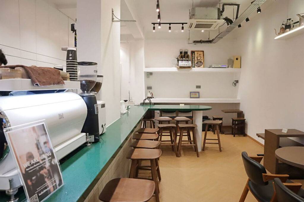 Minimalist coffee shop interior with a long green bar counter, wooden stools, and simple white walls. Espresso grinder visible.