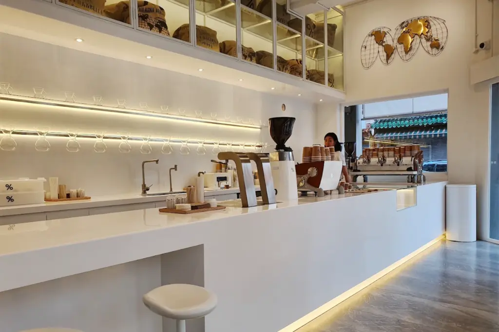 Interior of the %Arabica coffee shop in Arab Street, showcasing a minimalist white aesthetic with a long service counter, professional espresso equipment, glass-enclosed coffee bean storage, and a decorative gold world map.