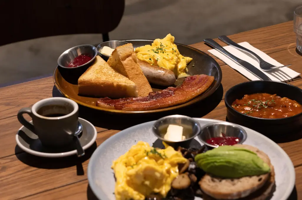 Two plates of hearty breakfast food on a wooden table. The main plate features scrambled eggs, toast, bacon, and a sausage, with a cup of black coffee and a side of baked beans.