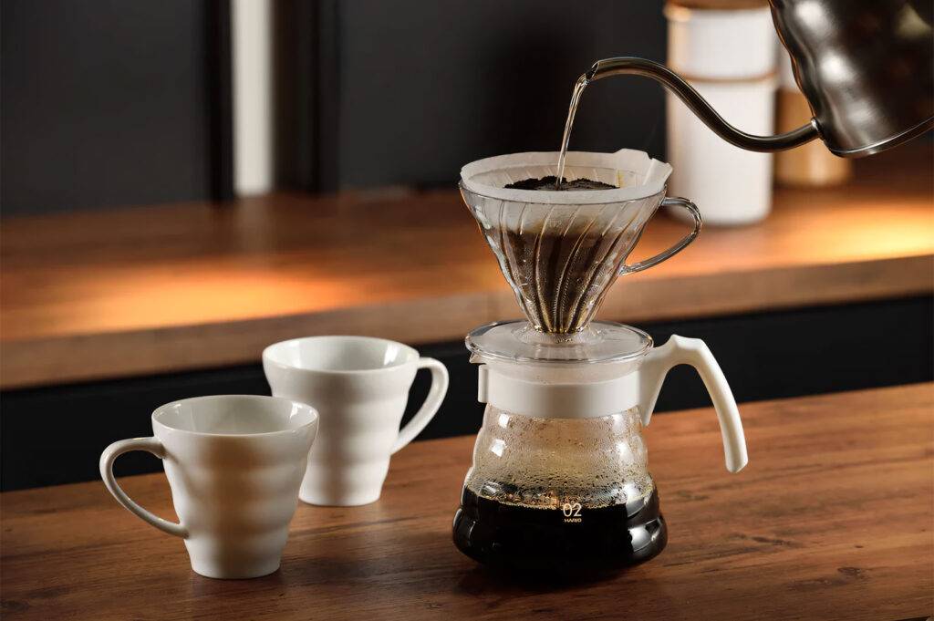 Close-up of a hand pouring hot water from a gooseneck kettle over coffee grounds in a Hario V60 dripper on a wooden counter.