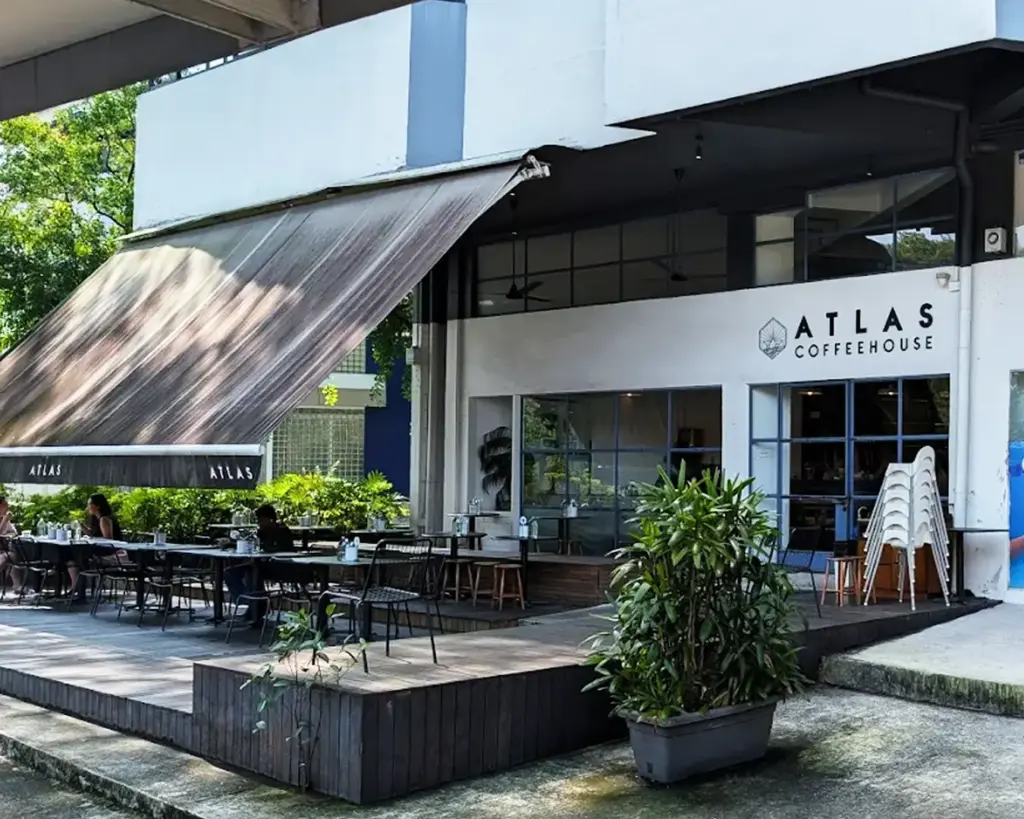 Outdoor view of Atlas Coffeehouse featuring a shaded patio with tables and chairs. The facade is modern, surrounded by greenery, creating a relaxed ambiance.