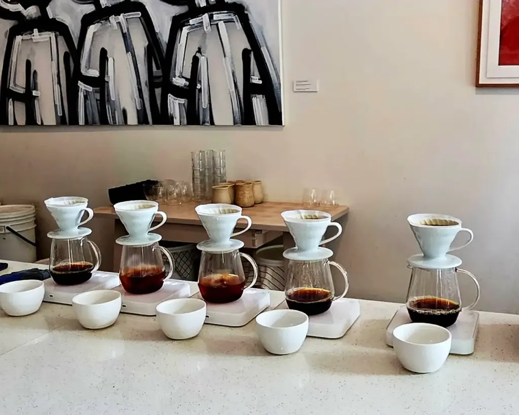 Four pour-over coffee makers line a countertop, paired with white cups, against a backdrop of abstract black and white art, creating a calm, artistic setting.