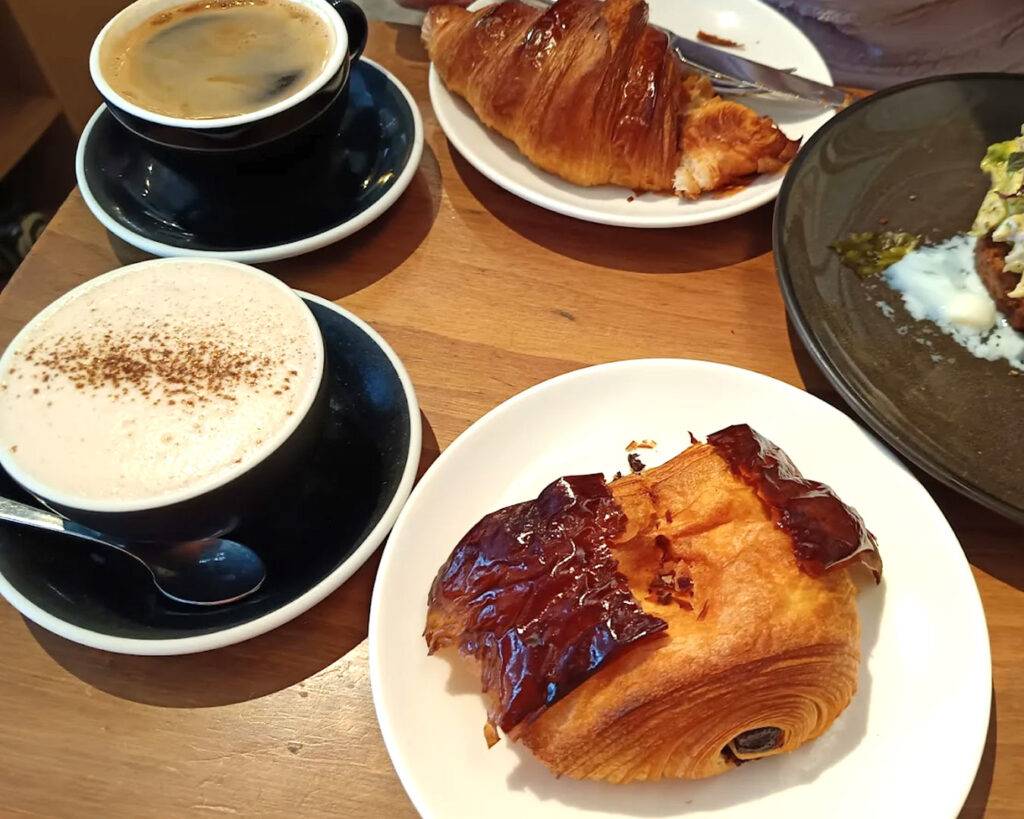 A table set with freshly brewed coffee and an assortment of pastries inside an industrial-style café, featuring exposed brick walls and metal fixtures that reflect the café’s heritage-inspired design.