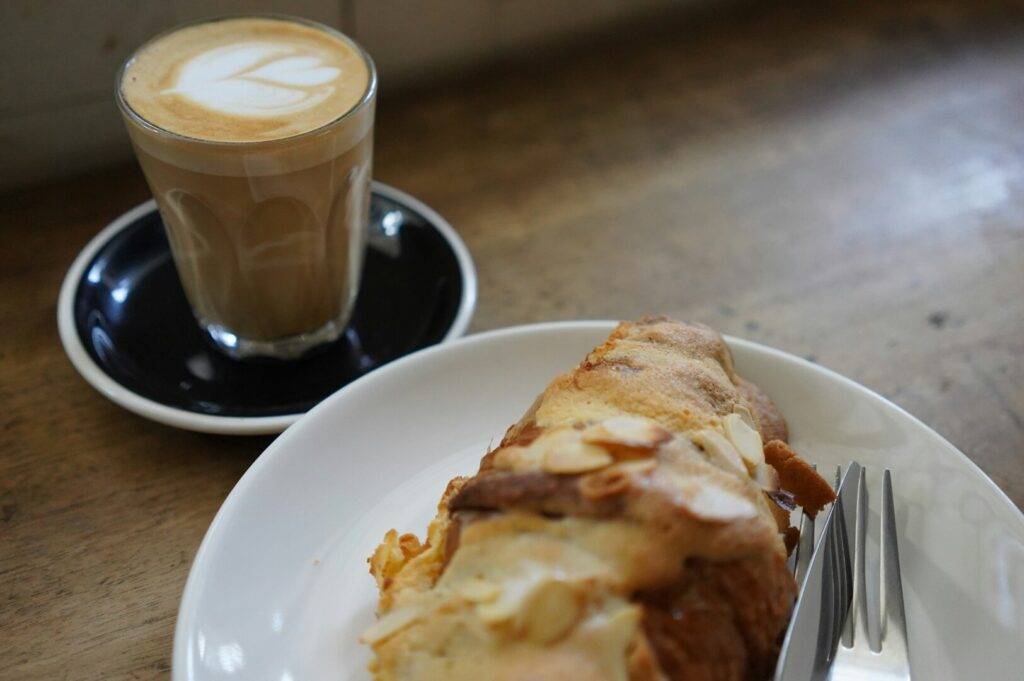 A latte with heart-shaped latte art in a glass on a black saucer sits beside a plate with a flaky almond croissant on a rustic wooden table. Cozy ambiance.