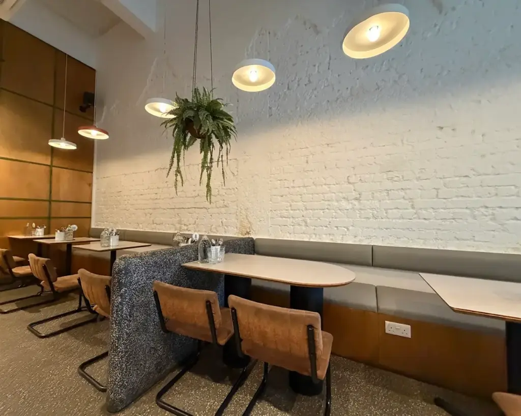 Chic café interior with wooden tables, brown chairs, hanging ferns, and warm pendant lights. Textured white brick wall adds a cozy atmosphere.