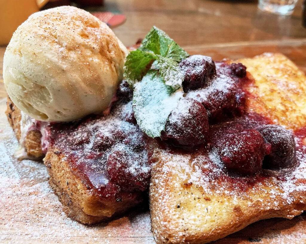 A golden brioche French toast topped with a scoop of ice cream and light syrup drizzle, served on a ceramic plate with soft natural lighting highlighting the texture and sweetness of the dessert.