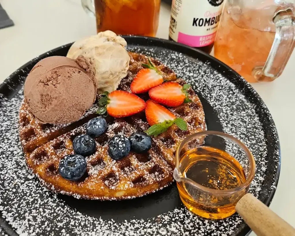 A warm waffle topped with a scoop of creamy ice cream and a mix of fresh berries, with syrup served on the side. The dish combines sweet, fruity, and indulgent flavours with contrasting textures.