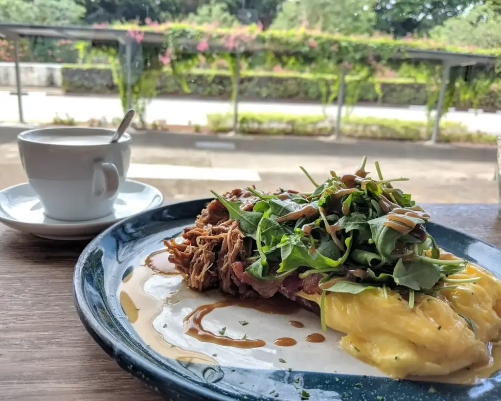 Plate with arugula and sauce, alongside creamy scrambled eggs. A cup of coffee sits beside, with a garden view outside.