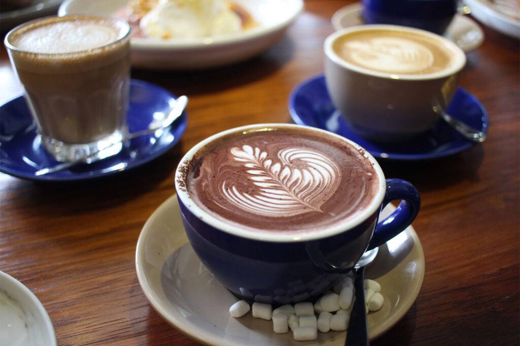 Close-up of three coffee drinks on a wooden table, featuring a dark blue cup of hot chocolate with detailed leaf latte art and mini marshmallows.