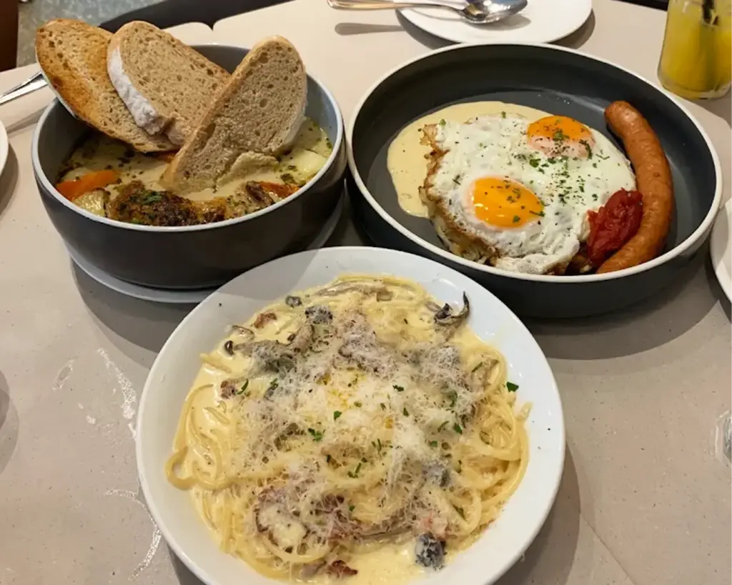 Three dishes on a table: creamy pasta with mushrooms, poached eggs with sausage and tomato, and a stew with vegetables and slices of bread.