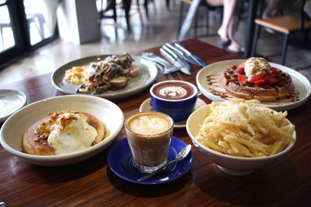 Overhead shot of a full brunch spread on a wooden table, including coffee, pancakes with ice cream, savory mushrooms on toast, a waffle with strawberries, and truffle fries.