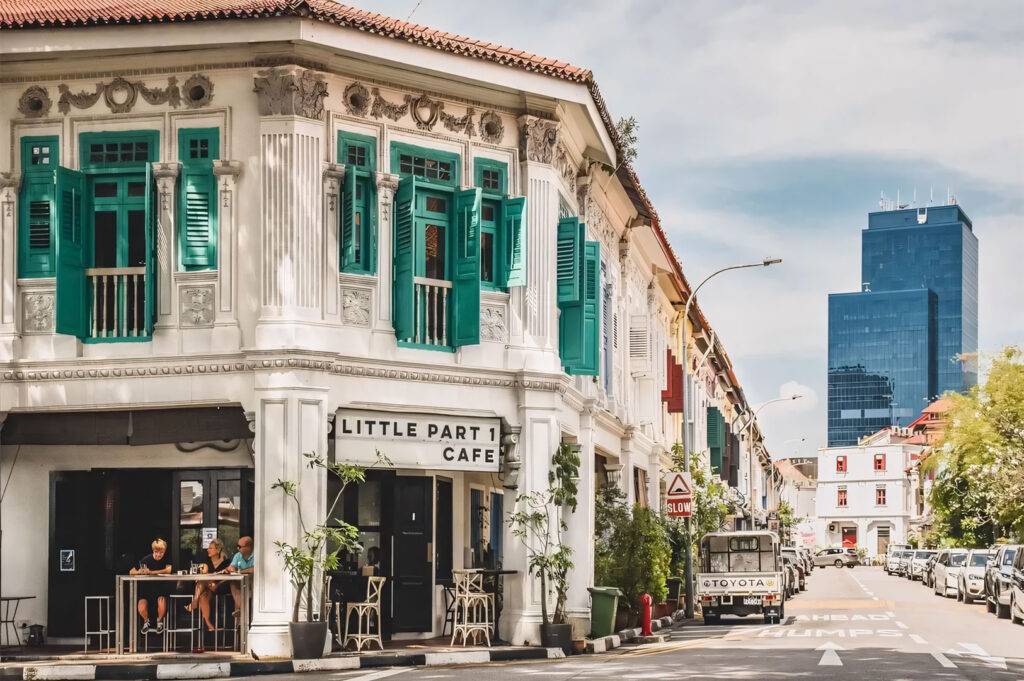 Exterior view of 'Little Part 1 Cafe' in a traditional white shophouse building with teal/green shutters in a sunny Singapore streetscape, with modern skyscrapers in the distance.