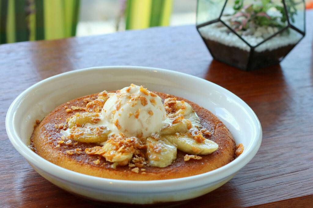 Close-up of a bowl of fluffy pancakes topped with sliced bananas, vanilla ice cream, and crunchy granola.