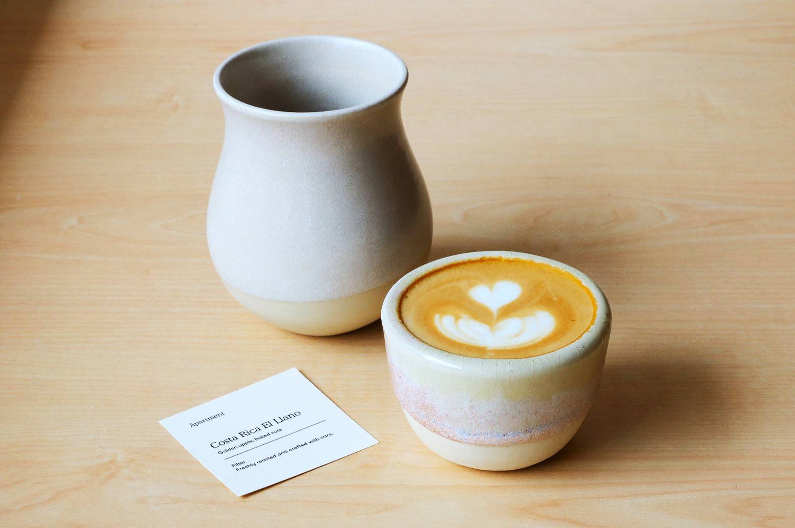 A latte with heart latte art in a ceramic cup sits on a wooden table alongside a matching vase and a card. The scene is calm and elegant.