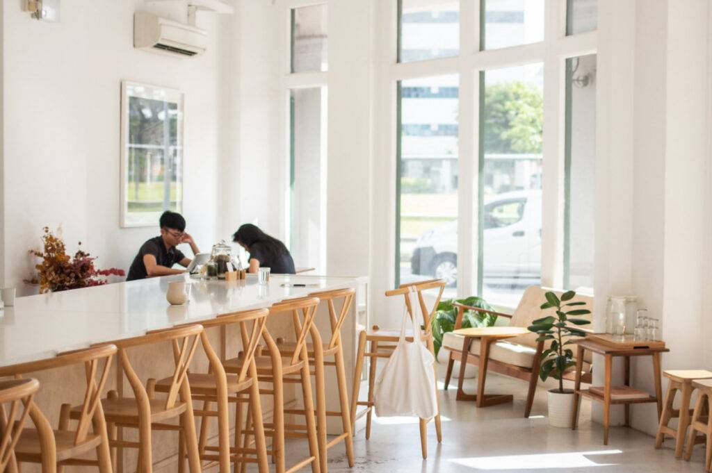 Bright, minimalist cafe with wooden chairs and plants. Two people sit at a long white counter, engaged in conversation. Large windows bring in natural light.