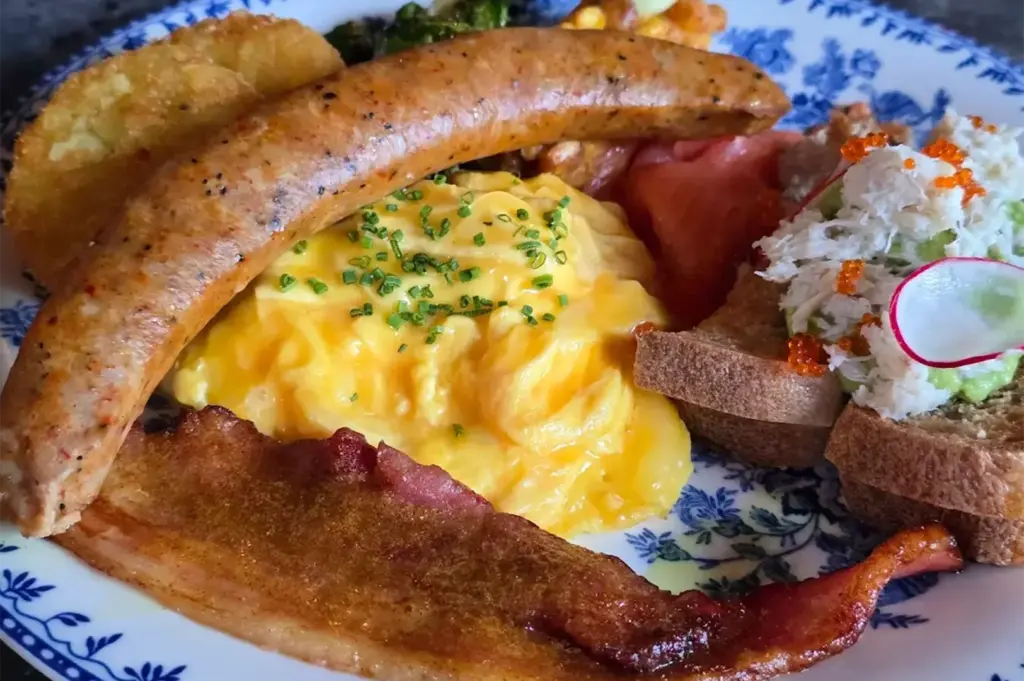 A hearty breakfast plate featuring juicy sausages, fluffy scrambled eggs, crispy hash browns, and strips of golden-brown bacon, served warm and freshly cooked at Symmetry in Bugis.