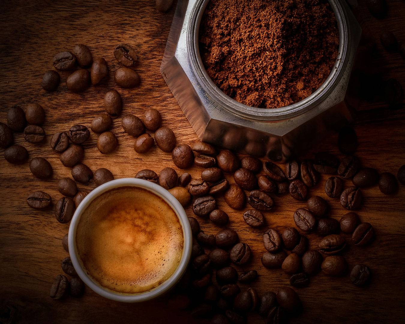 A close-up of roasted coffee beans scattered beside a freshly brewed cup of black coffee, with light steam rising to show warmth and freshness.