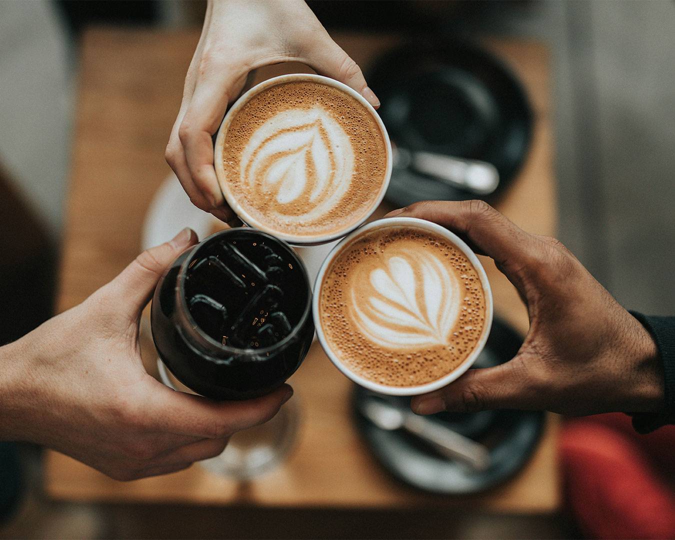 Three different hands holding takeaway cups of coffee together, captured in a close-up shot that highlights warmth, connection, and a shared coffee moment.