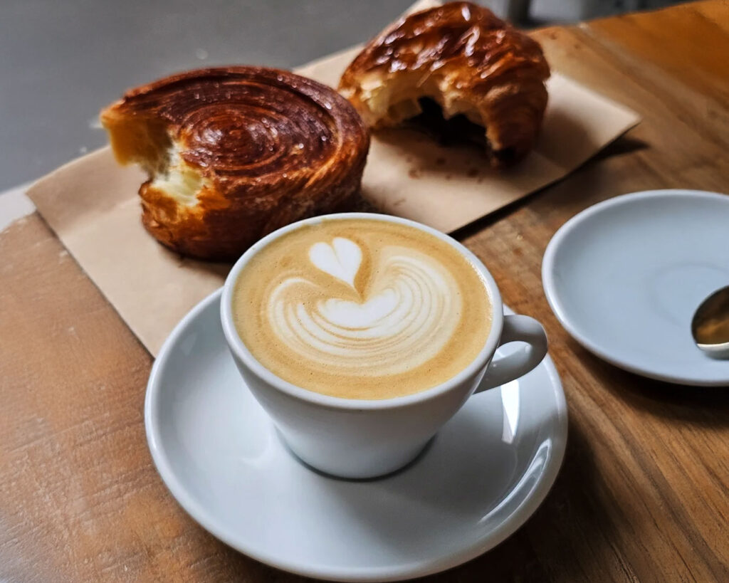 A cup of freshly brewed coffee served alongside golden pastries on a café table at Nylon Coffee Roasters, with natural light highlighting the warm tones of the drinks and baked goods for a cosy breakfast scene.