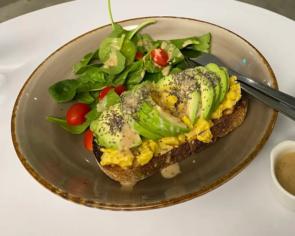 Avocado toast topped with sliced avocado, scrambled eggs, and chia seeds, accompanied by a spinach and cherry tomato salad drizzled with dressing.