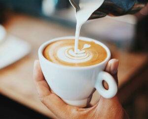 A close-up of a hand gently pouring fresh milk into a cup of hot coffee, creating smooth swirls of foam on the surface.