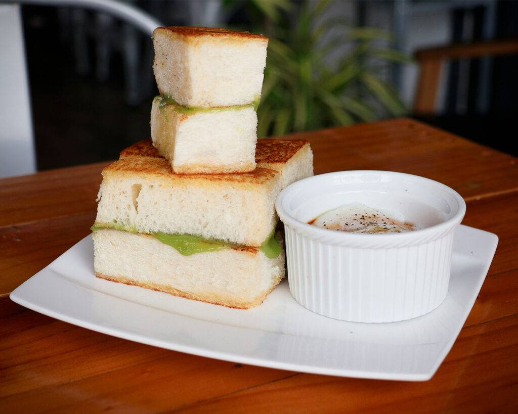 Two slices of golden-brown toasted bread filled with kaya and butter, served on a plate with a simple café setting in the background.