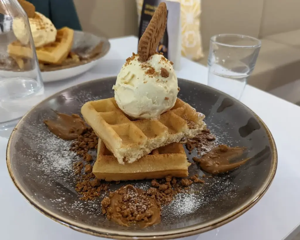 A plate of waffles topped with a scoop of vanilla ice cream and a cookie. Crumbled cookie bits and caramel sauce accent the dish.