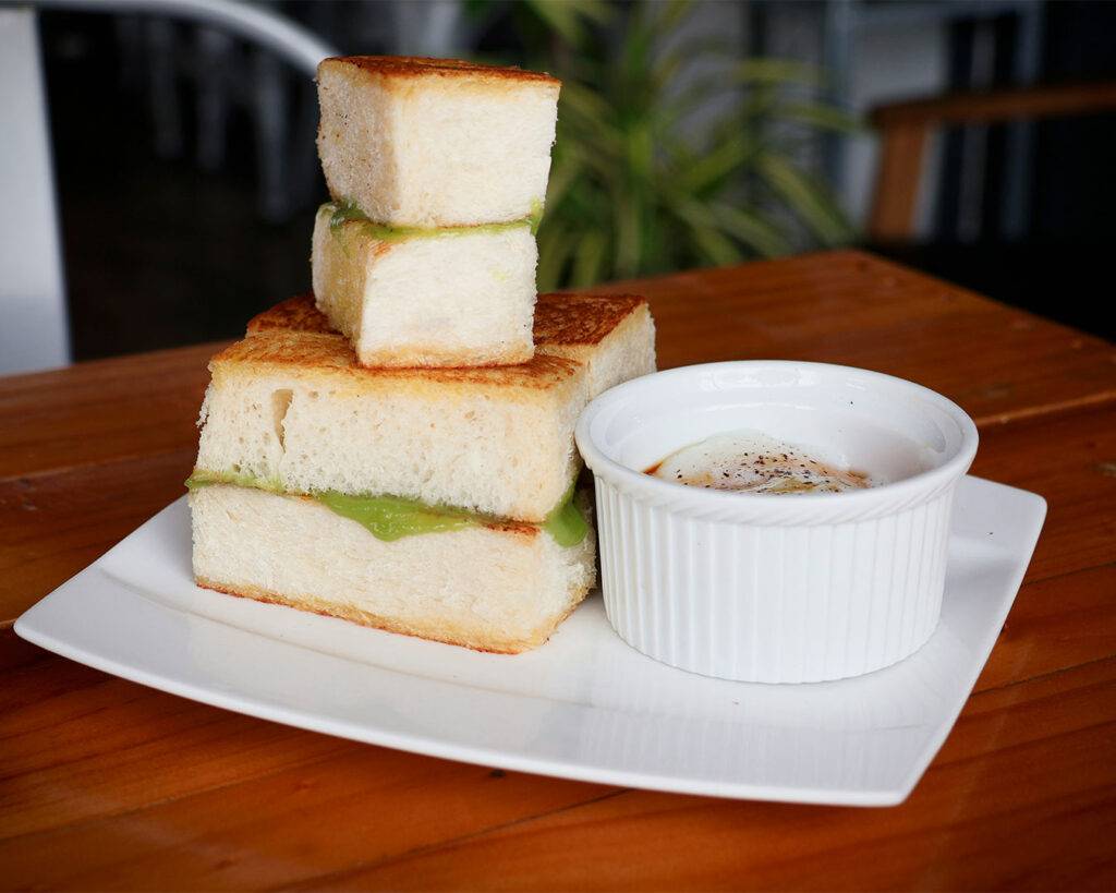 A traditional breakfast set of crispy kaya toast served with two soft-boiled eggs and a small bowl of soy sauce and pepper.