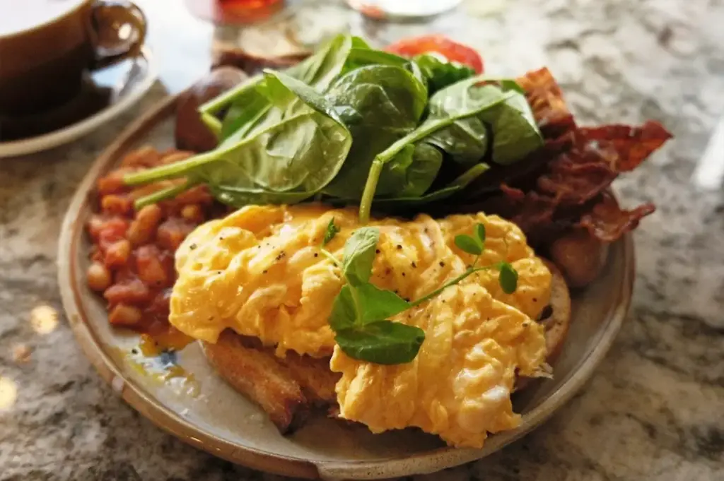 A wholesome breakfast plate of scrambled eggs, crispy hash browns, and bacon, complemented by a side of fresh green spinach for a balanced brunch at Common Man Coffee Roasters.