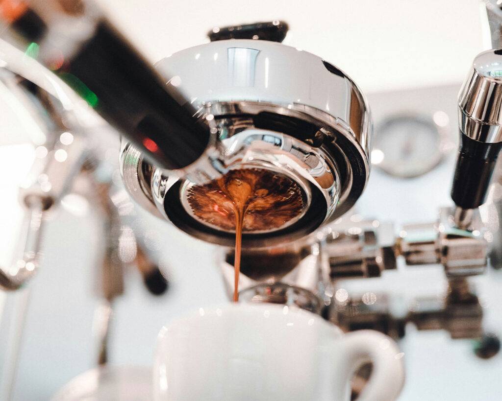 A stainless steel espresso machine with a portafilter attached, preparing coffee as hot water is extracted through finely ground coffee.