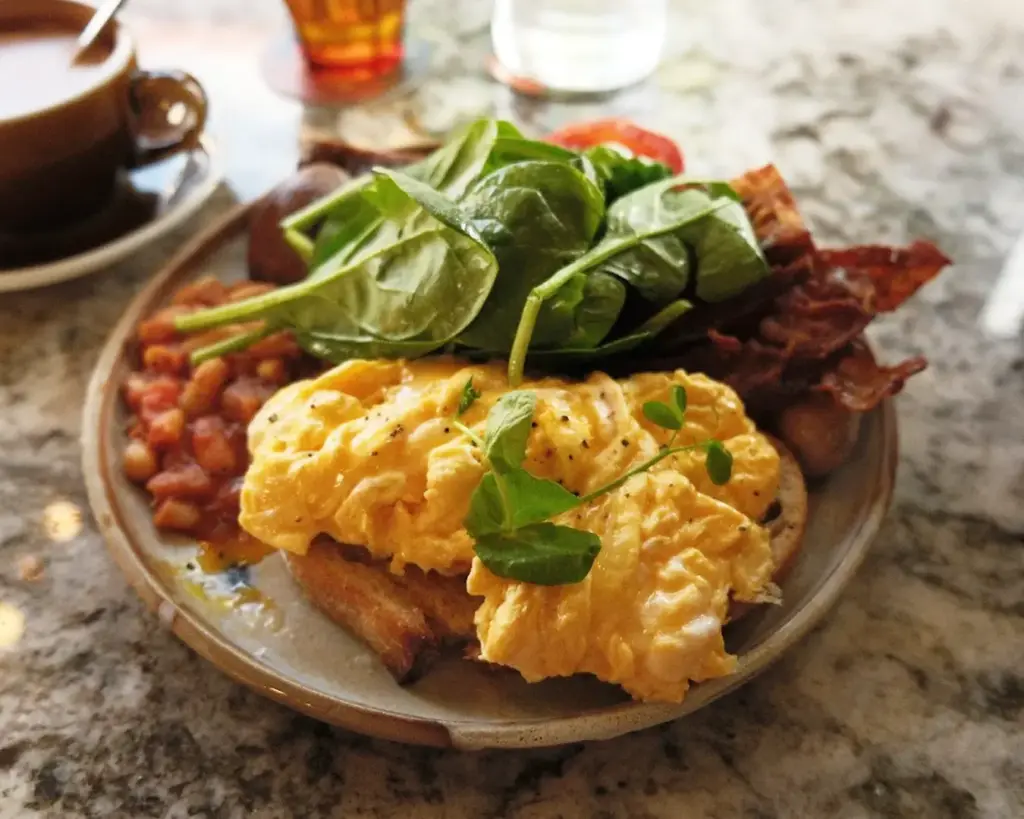 A wholesome breakfast plate of scrambled eggs, crispy hash browns, and bacon, complemented by a side of fresh green spinach for a balanced touch.