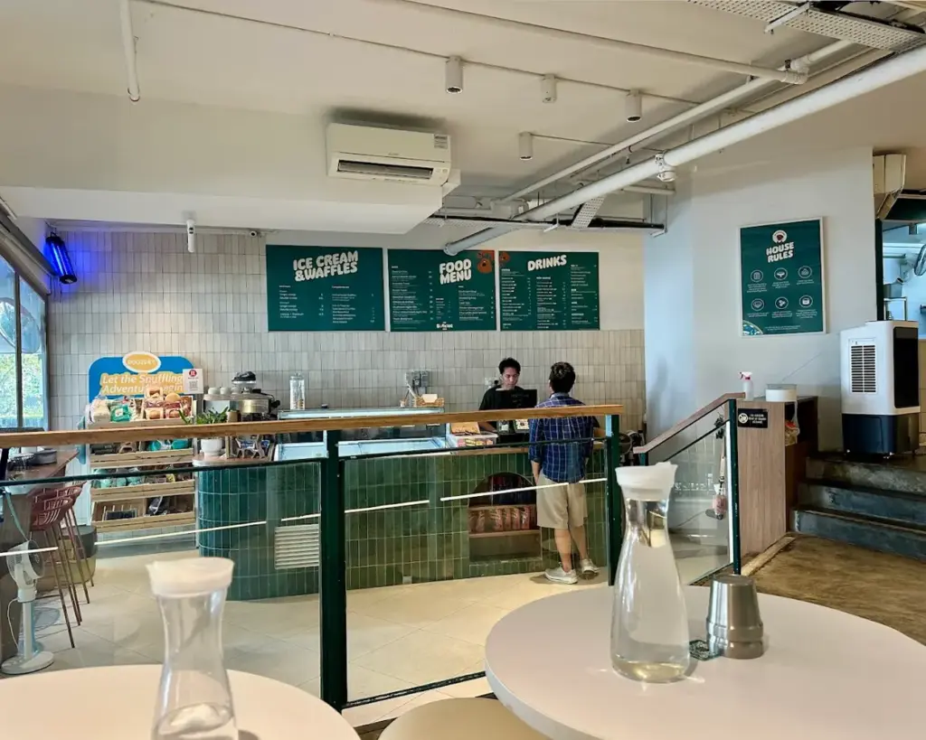 A cozy cafe interior with a counter at the center. Two people are at the counter under green menus labeled "Ice Cream & Waffles," "Food Menu," and "Drinks." The space is bright, with a modern feel, white walls, and glass bottles on round tables.