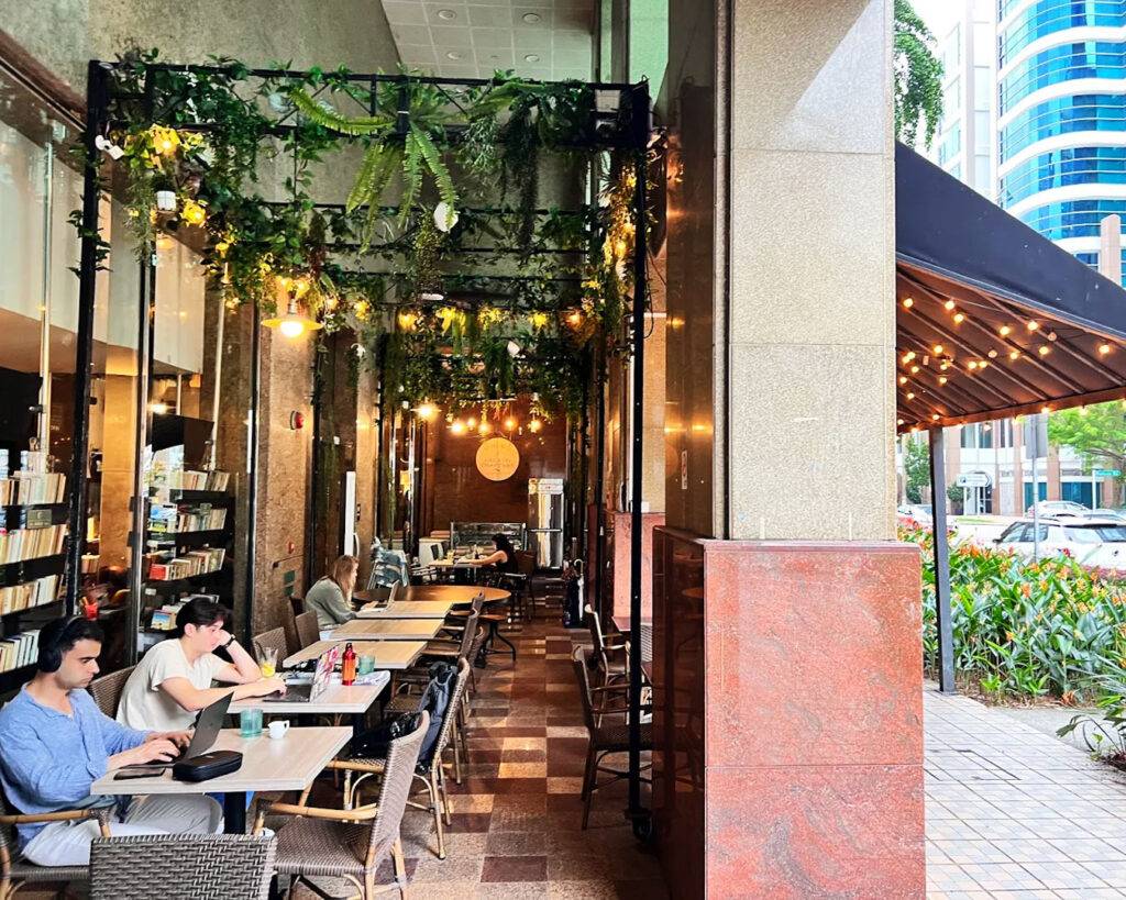 An outdoor dining space with tables and chairs set under natural light, surrounded by greenery, creating a relaxed and breezy environment for guests to enjoy meals beside the river.