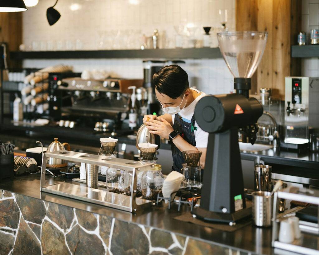 A barista carefully pouring hot coffee from a kettle into a cup behind the café counter, showing professional brewing technique.