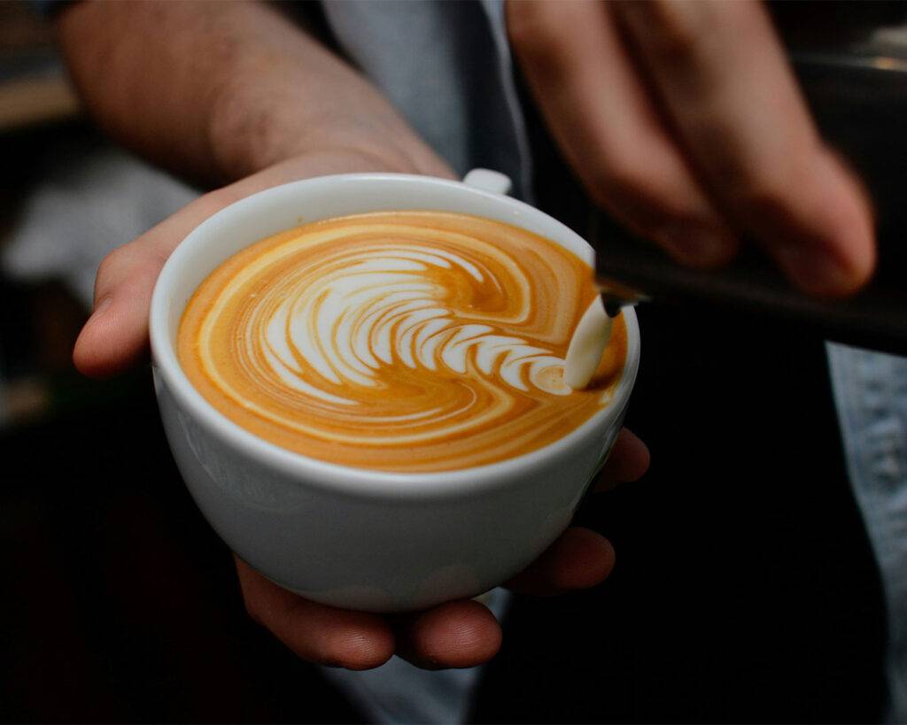 A barista carefully pouring steamed milk into a cup of coffee to create detailed latte art, demonstrating precision and craftsmanship.