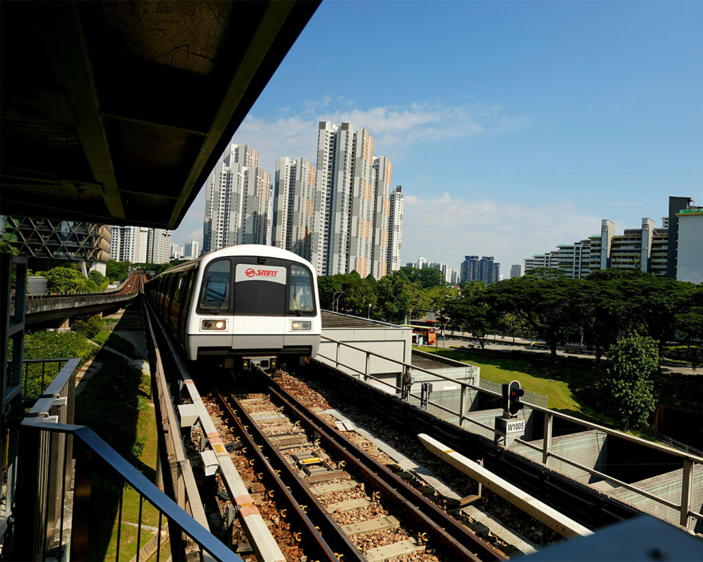 A Singapore MRT train arriving at an elevated station, with clean platforms, glass barriers, and city buildings visible in the background under a bright sky.