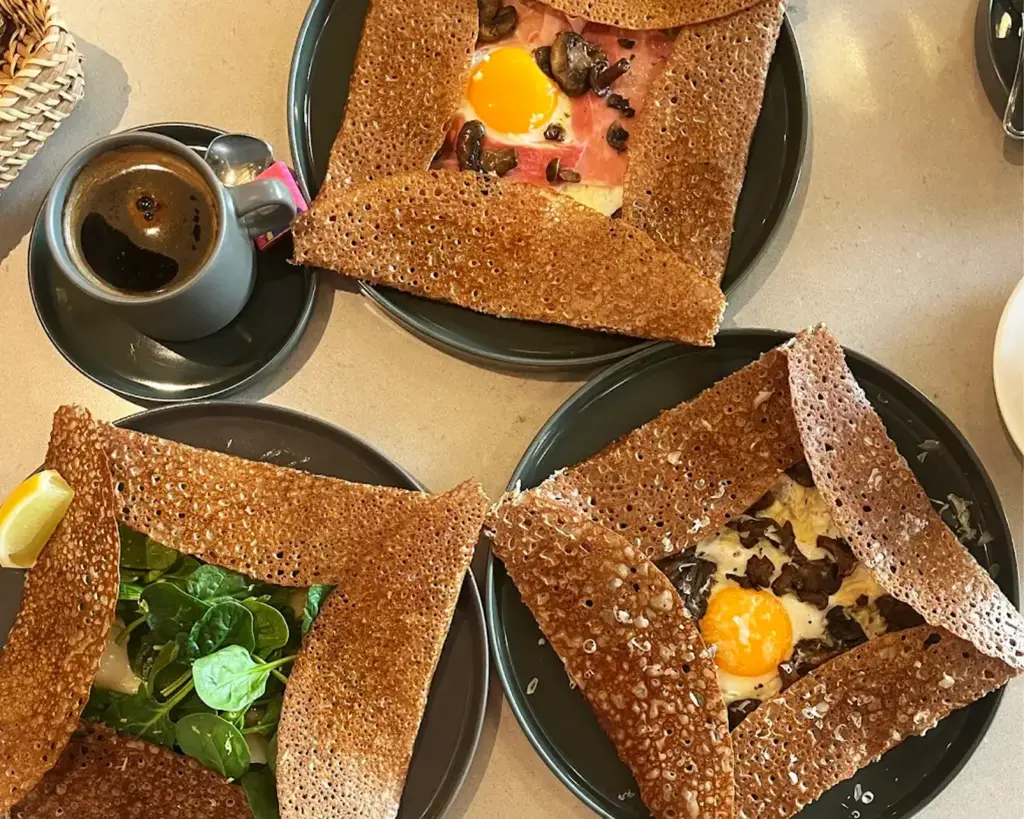 Three beautifully plated savoury galettes on a dining table, featuring crisp buckwheat crêpes filled with ingredients like eggs, cheese, and ham, presented in a warm and inviting brunch scene.