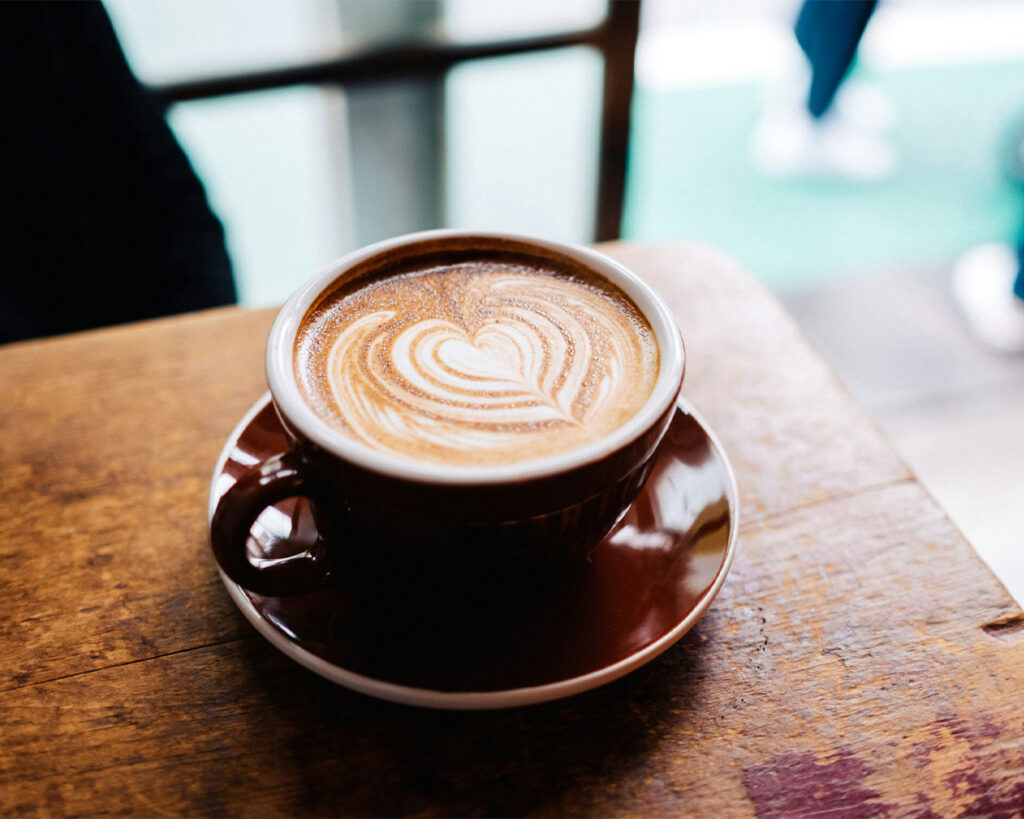 A cup of hot coffee topped with delicate latte art in a leaf pattern, placed on a saucer with natural light highlighting the creamy foam and rich coffee colour.