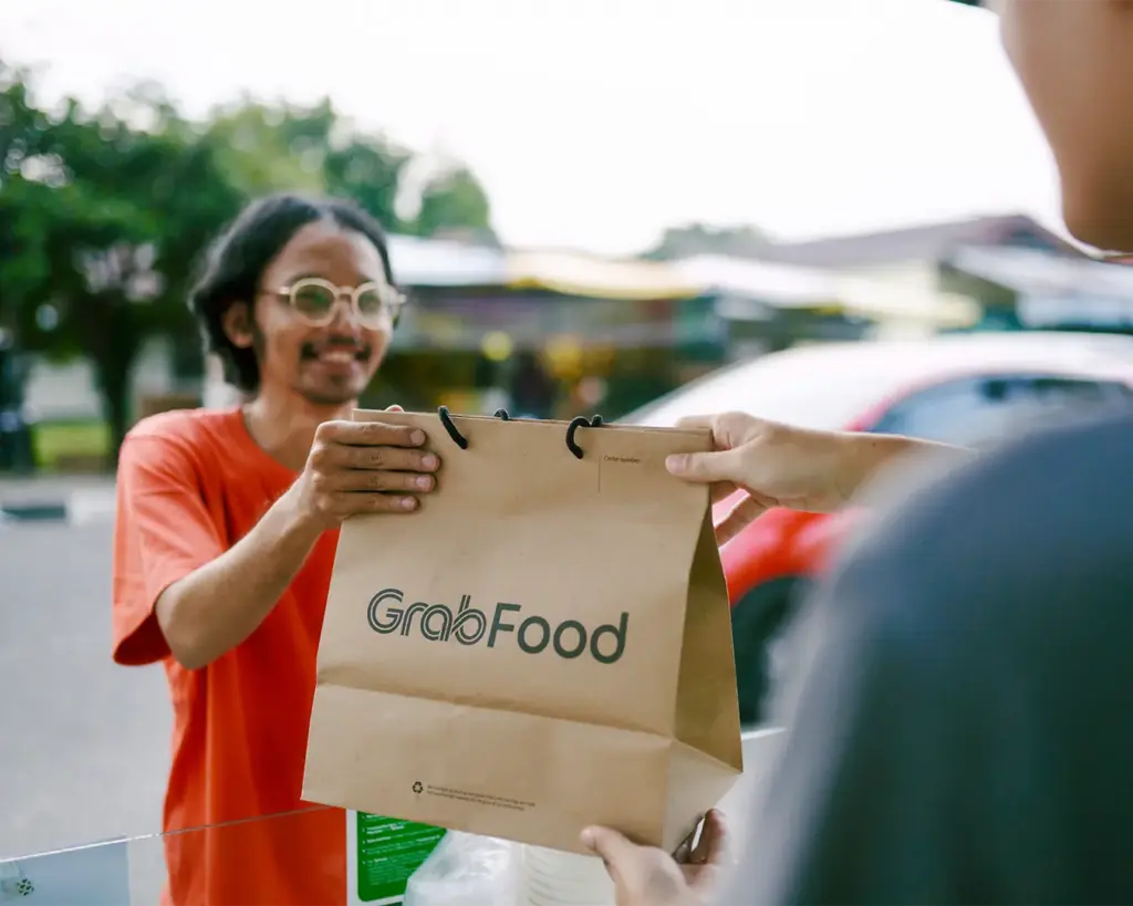 A person handing over a GrabFood takeaway bag, representing a food delivery order being collected or delivered, symbolising the convenience of ordering meals through delivery services.