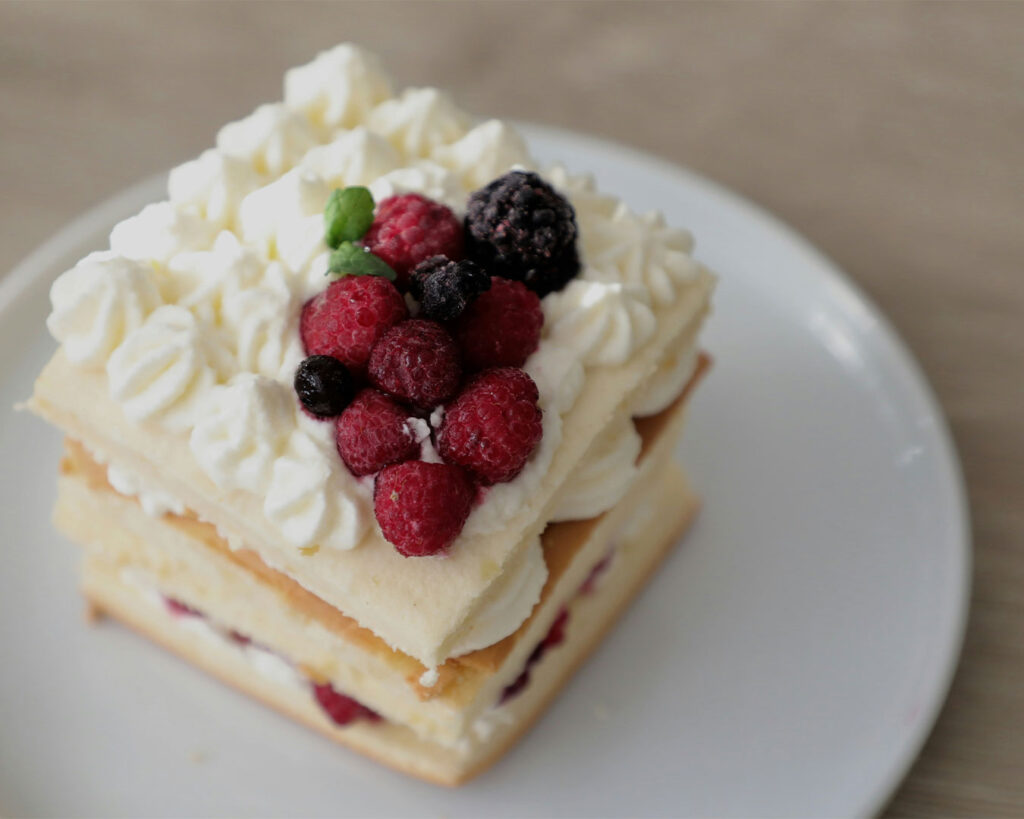 A slice of dessert cake topped with fresh berries such as strawberries and blueberries, finished with a light dusting of icing sugar and served on a clean plate.