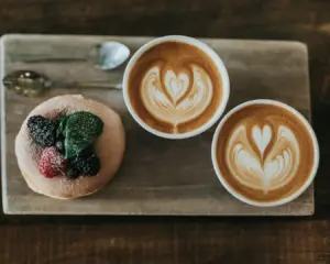 Two cappuccinos with heart latte art sit beside a donut topped with berries and mint on a wooden board, creating a cozy, inviting setup.