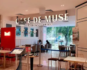 Interior of "Cafe de Muse," a dessert bar with cozy seating. Customers are seated near large windows, creating a bright, inviting atmosphere.